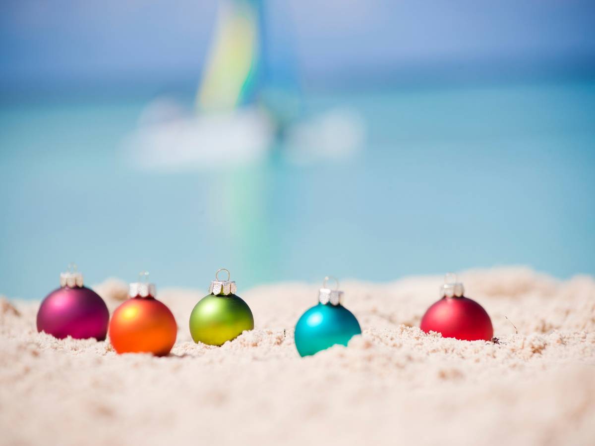 Colorful Christmas tree balls sitting in white sand with turquoise water and a sailboat in the background. 
