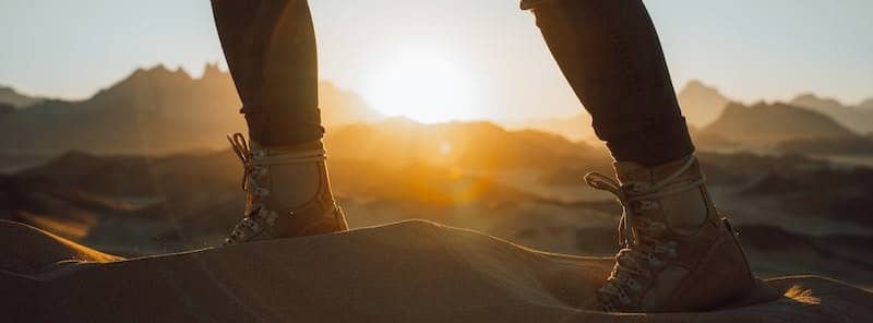 close up shot of person walking with his boots on a sand and sunshine at the far.