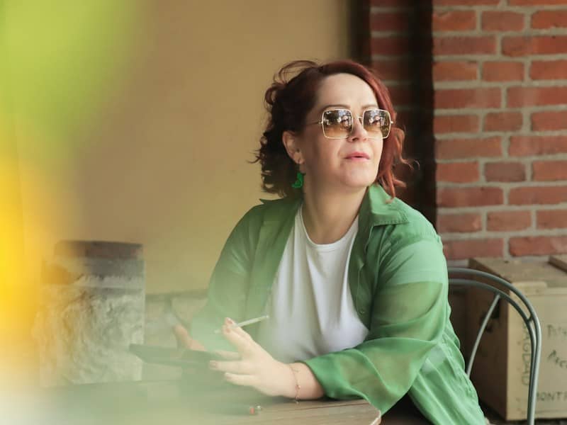 A woman with red hair wearing sunglasses and a green shirt holding a cigarette