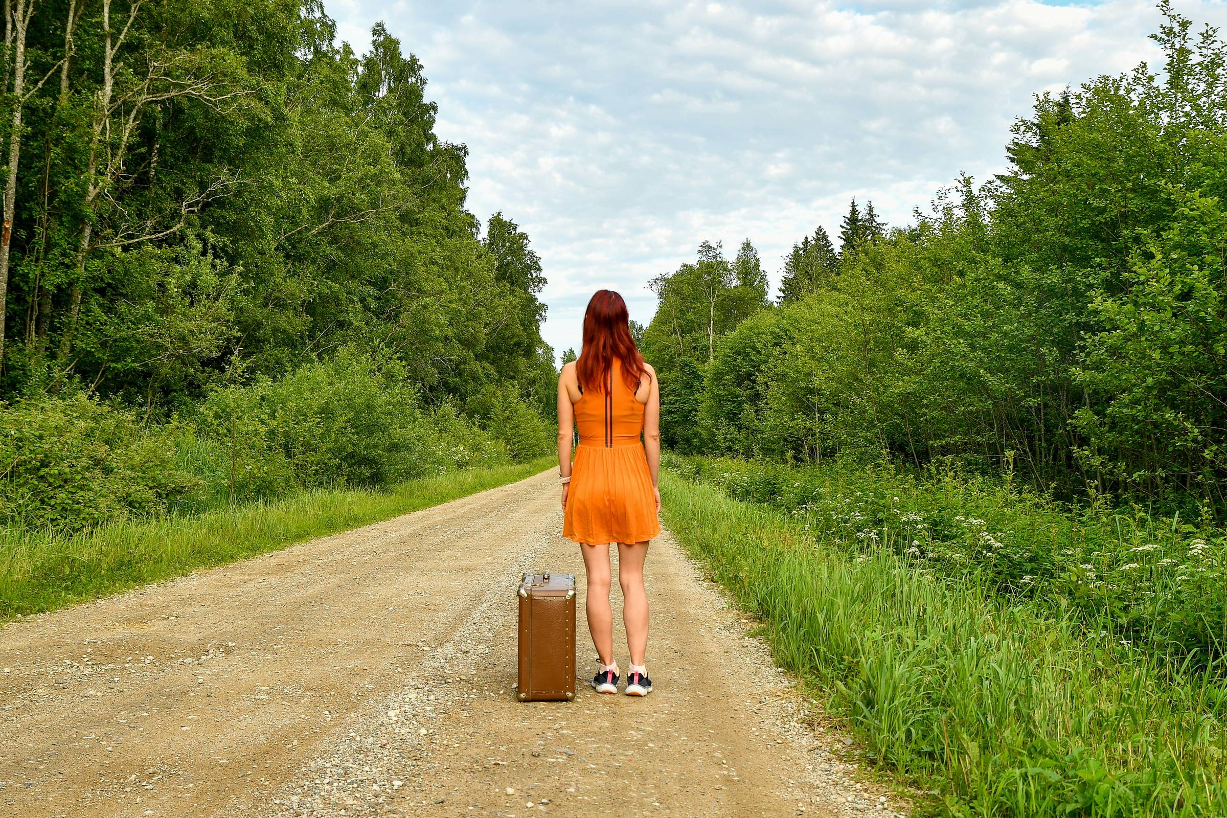A young red-haired woman in a bright orange dress standing on an empty gravel road with a vintage suitcase, hitchhiking towards a new adventure. 📸 Photo by Margo Evardson, IG: @margoevardson