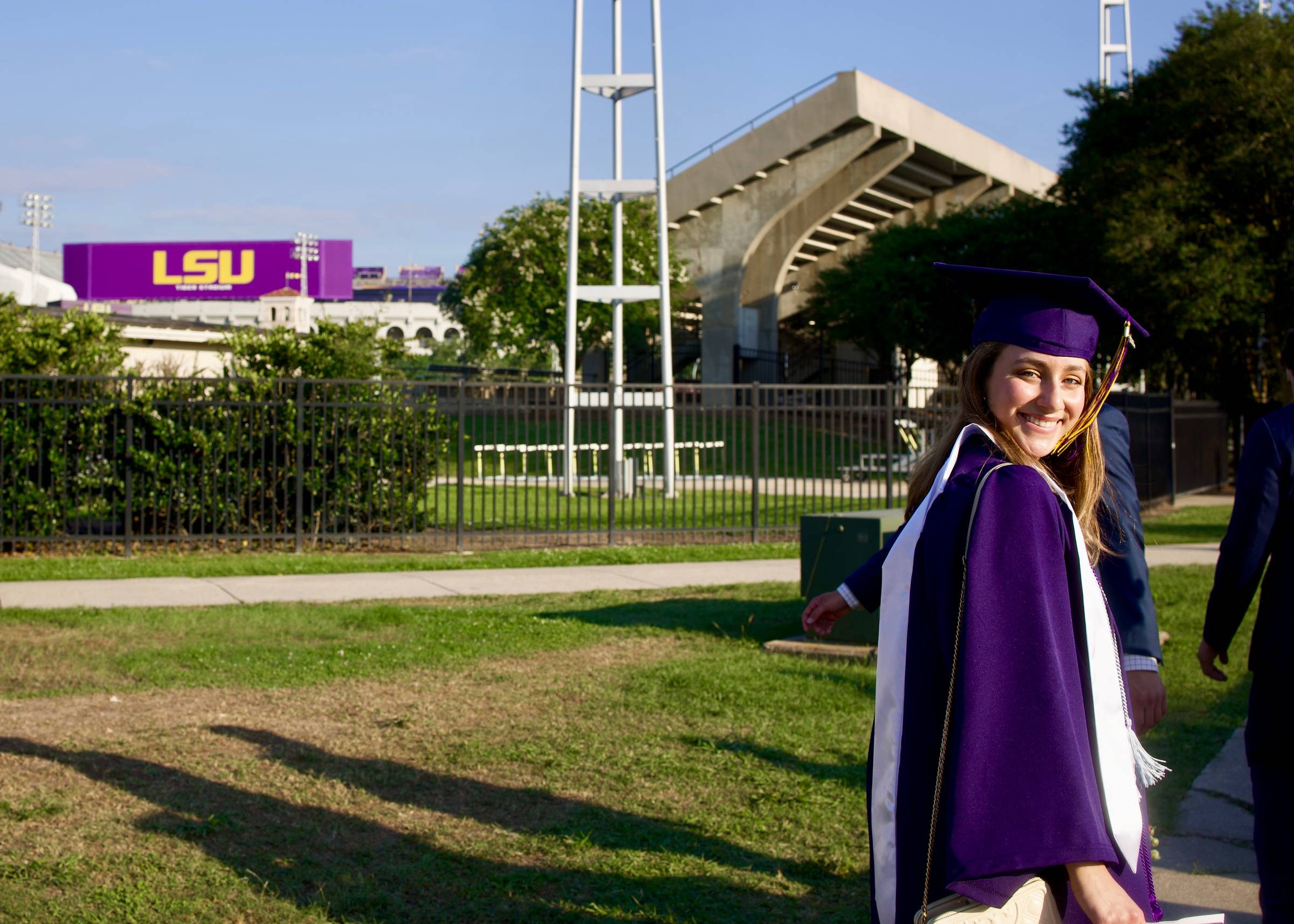 Grace Richard in LSU graduation cap and gown with stole, smiling, standing on the lawn outside Tiger Stadium.