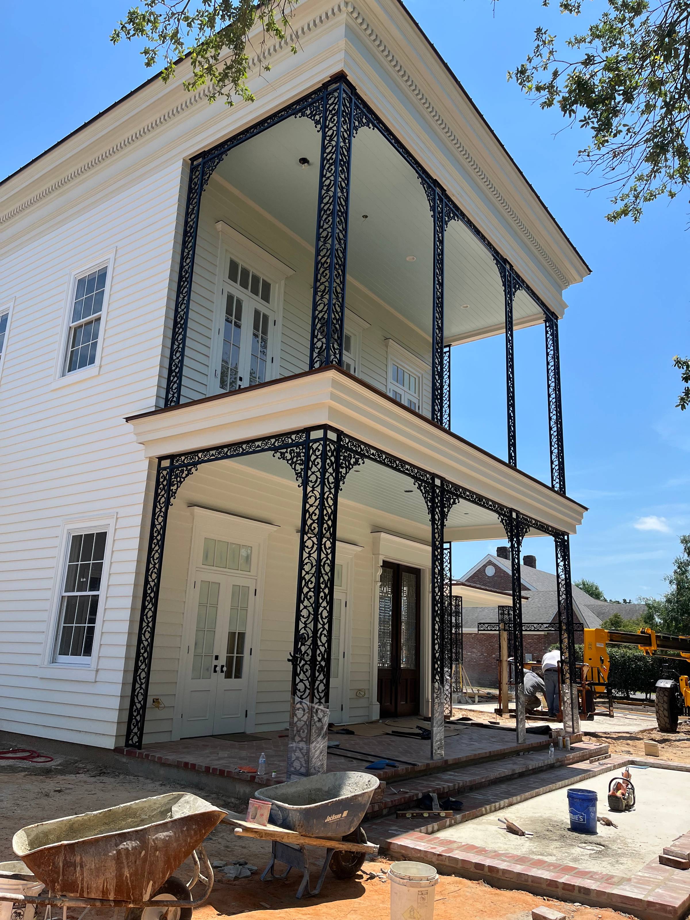 Two-story raised home under construction in West Monroe, Louisiana, with stacked galleries wrapped in black St. Charles-style ornamental ironwork against white siding.