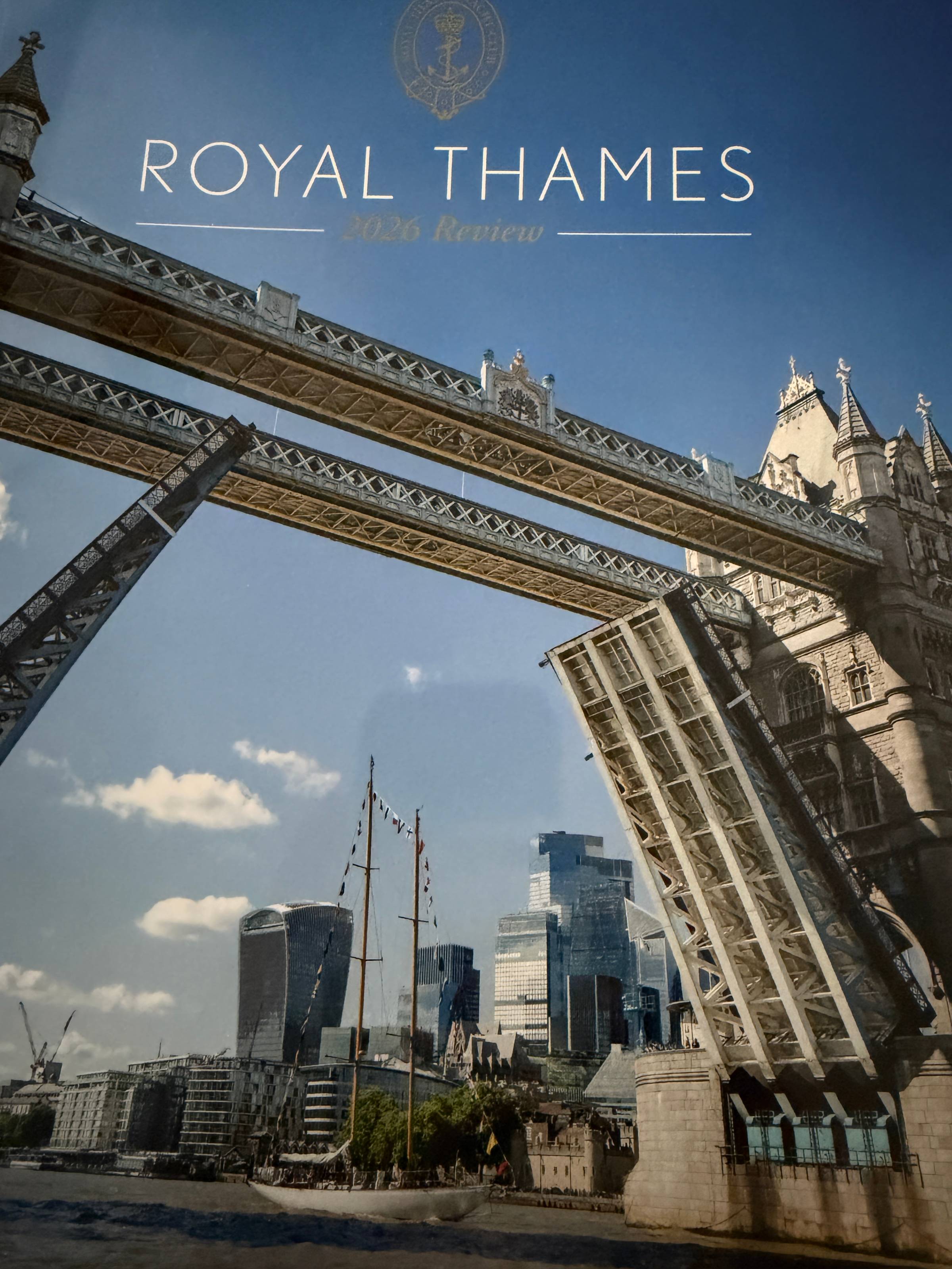 Cover of the Royal Thames Yacht Club's 2026 Review, showing Tower Bridge raised in London with a flagged sailing yacht passing through.