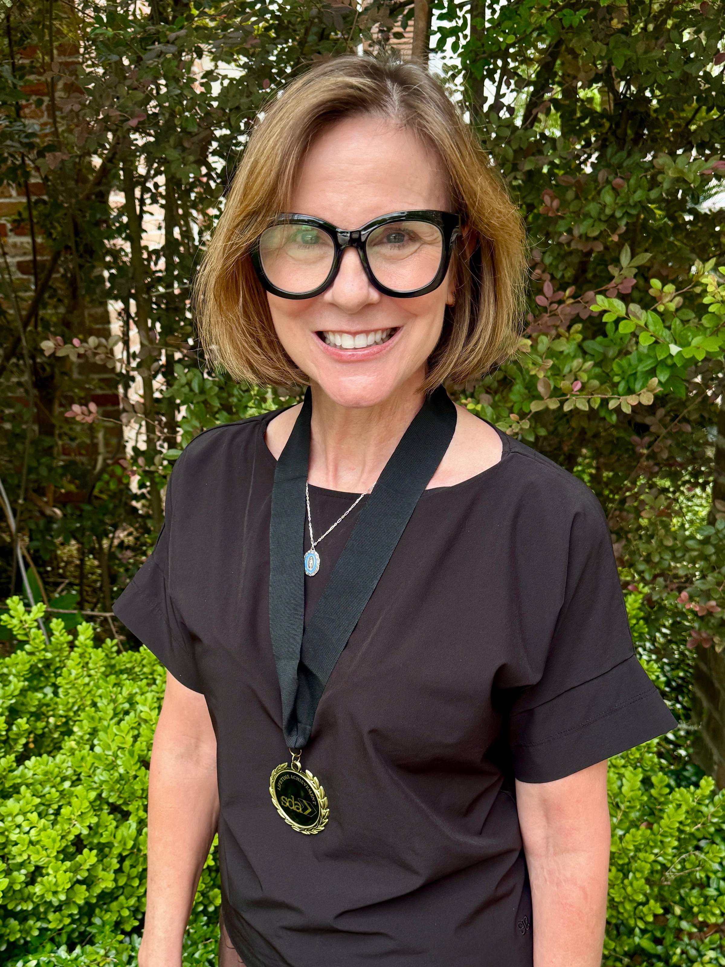 Liz Harris wearing the SDA Lifetime Achievement Award medal on a black ribbon, smiling outdoors with greenery in the background.