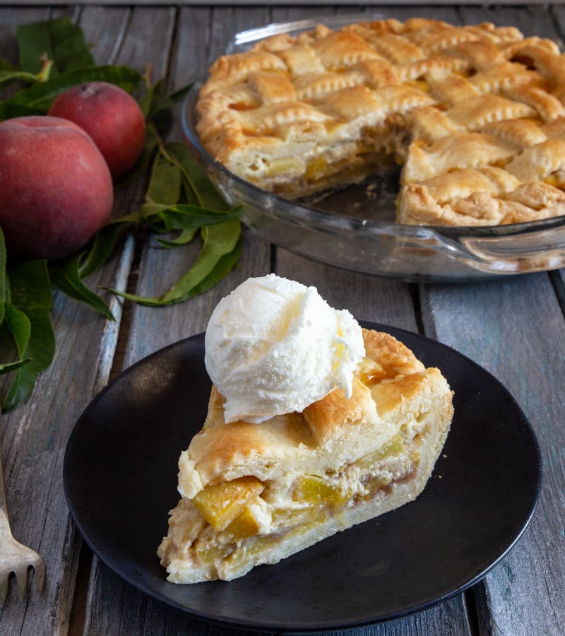 Peach pie in a glass dish and a slice with a scoop of ice cream on a black plate.