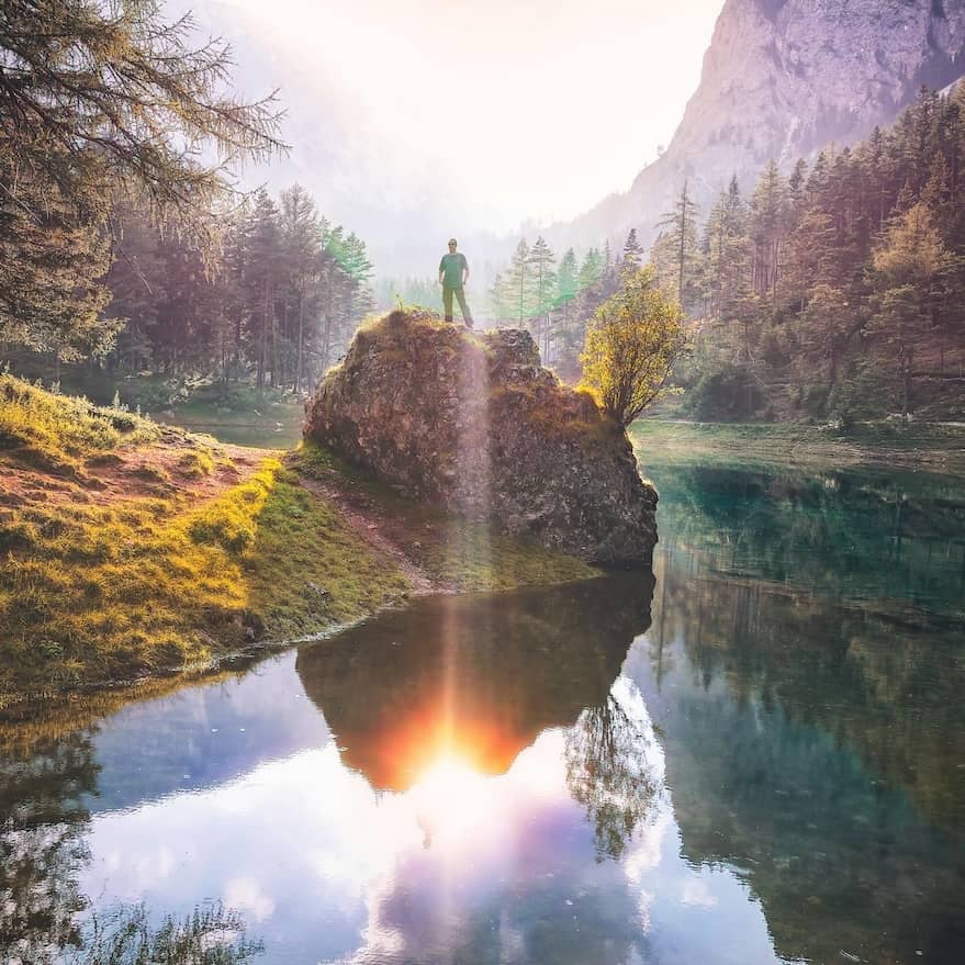 A person stands atop a large rock near a tranquil lake surrounded by lush greenery and dense trees. Sunlight streams down, creating a glowing reflection on the water. Misty mountains rise in the background, adding to the serene atmosphere.