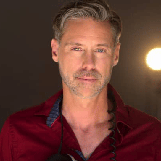 with a soft spotlight behind him, Roy, with some scruff, looks at the camera, wearing a button down maroon shirt.