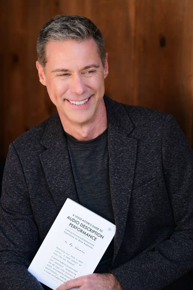 Glancing to the side with a smile, Roy holds his book A Voice Actor's Guide to Audio Description Performance.