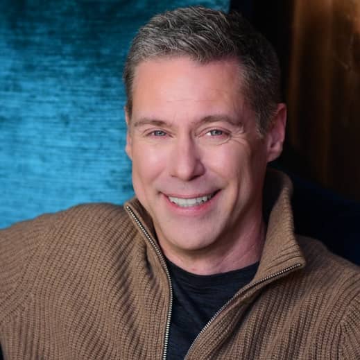 in front of an aqua colored fabric, Roy smiles at the camera, wearing a light brown zippered sweater.