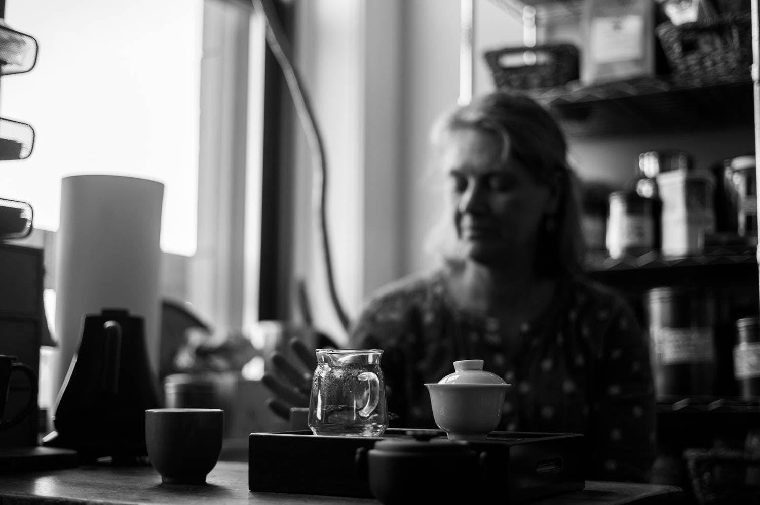 Black and white photo of a gaiwan and gong dao bei in the foreground and an out-of-focus woman drinking tea in the background 