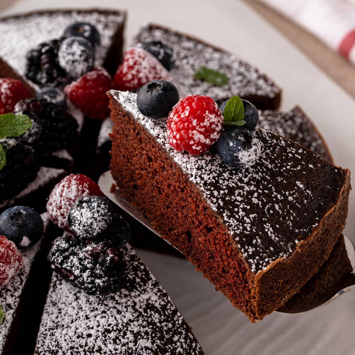 Cake server lifting up a slice of eggless chocolate cake that is dusted with powdered sugar and topped with fresh berries.