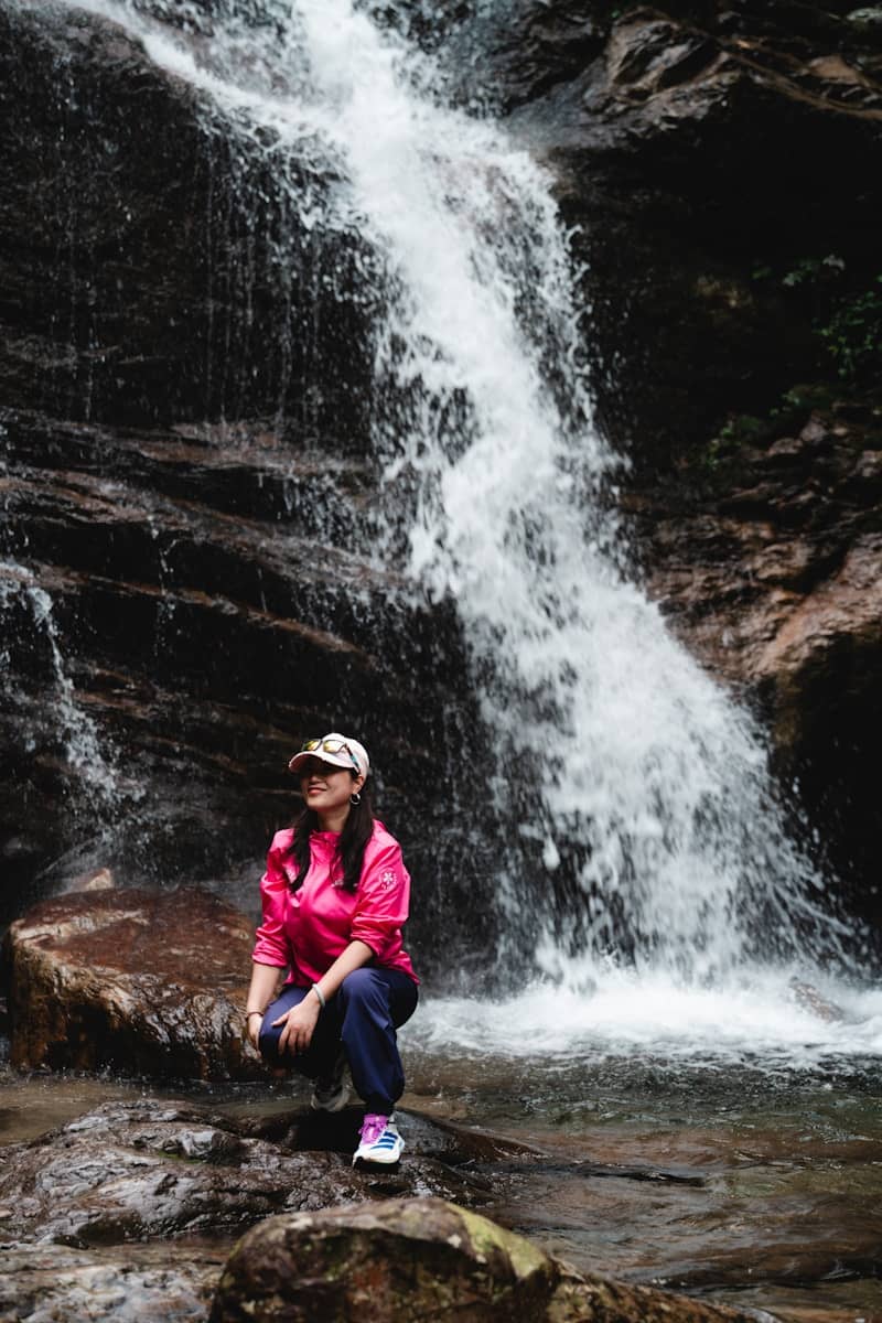 A woman in pink jacket crouching near a powerful waterfall.
