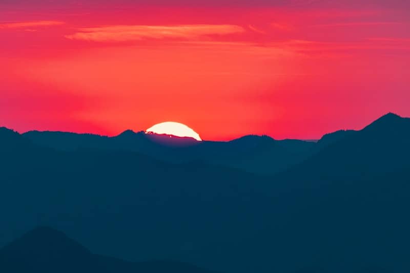A bright white sun disc behind dark blue mountain silhouettes under a red sky
