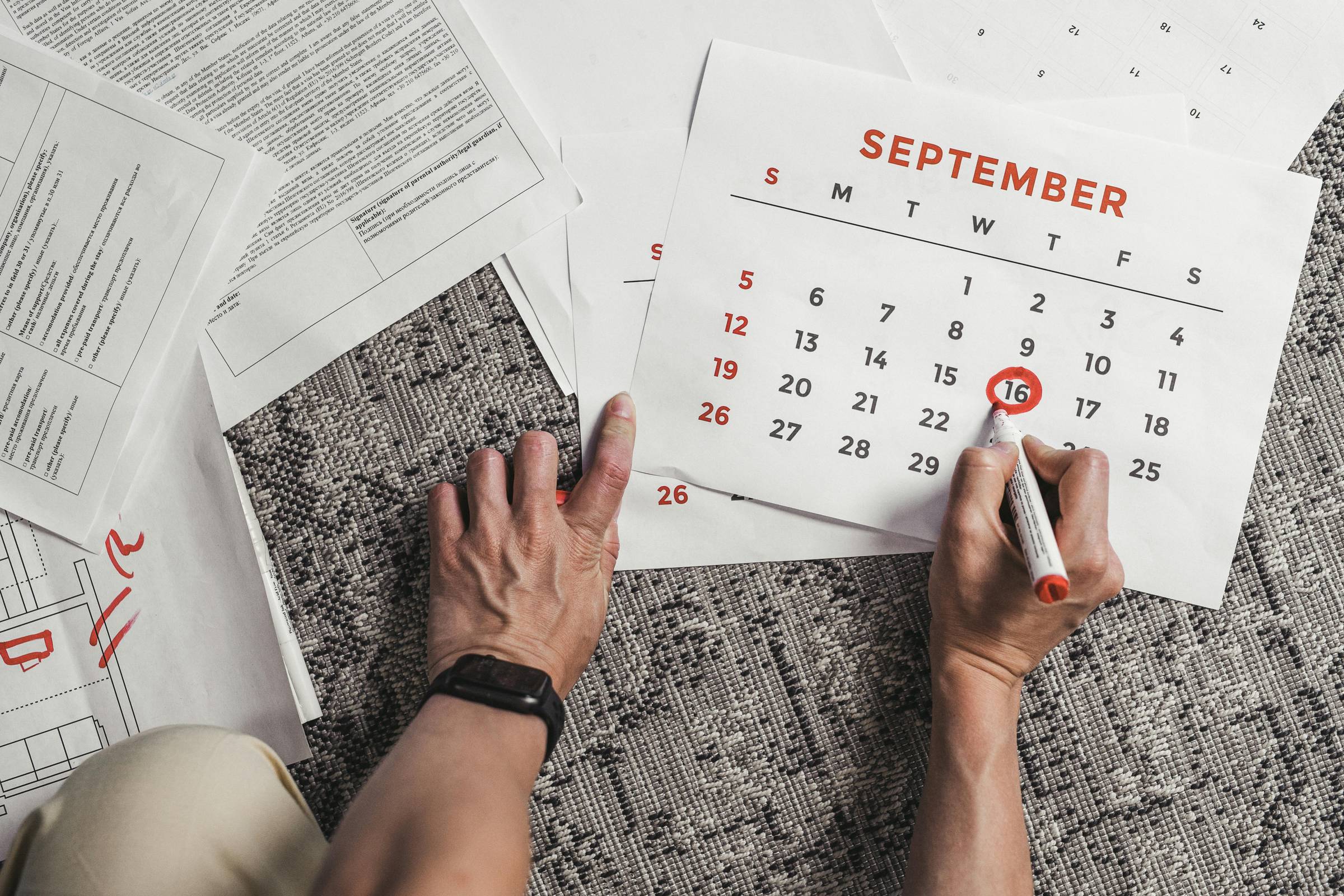 A person marking September 16 on a paper calendar with a red pen, surrounded by documents on a desk.