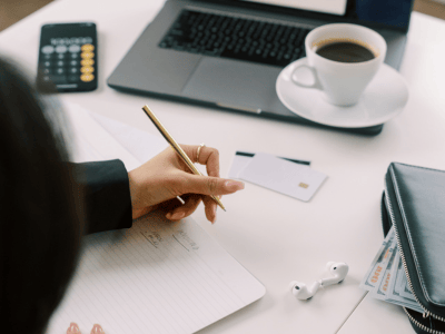 Woman taking notes at her desk with a laptop, cup of coffee, ear buds, calculator and wallet.