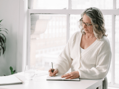 Woman journaling at a desk with water glass, focusing on creating a Stop-Doing List for business simplification and clarity.