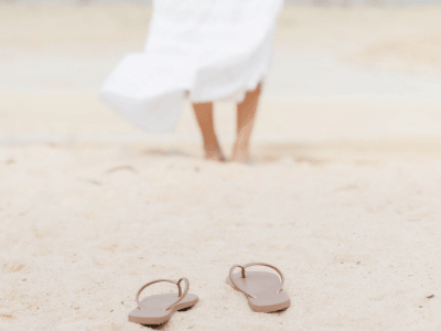Woman in white dress walking barefoot on the beach, sandals left in sand—symbolizing simplicity, calm, and sustainable business energy.