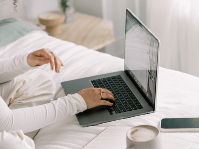 Woman typing on a laptop while sitting on a bed with coffee and phone nearby, representing blogging and online work in 2025.