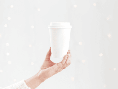 A minimalist and aesthetic image of a hand holding a plain white coffee cup against a softly blurred background with twinkling lights, evoking a cozy and simple offer.