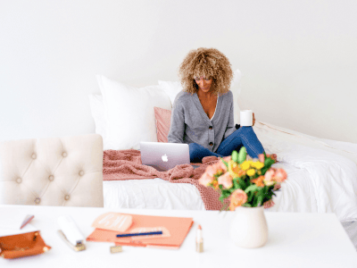 Woman sitting on bed with laptop and coffee, reviewing business offers in a calm workspace; flowers and planner suggest aligned, intentional strategy.