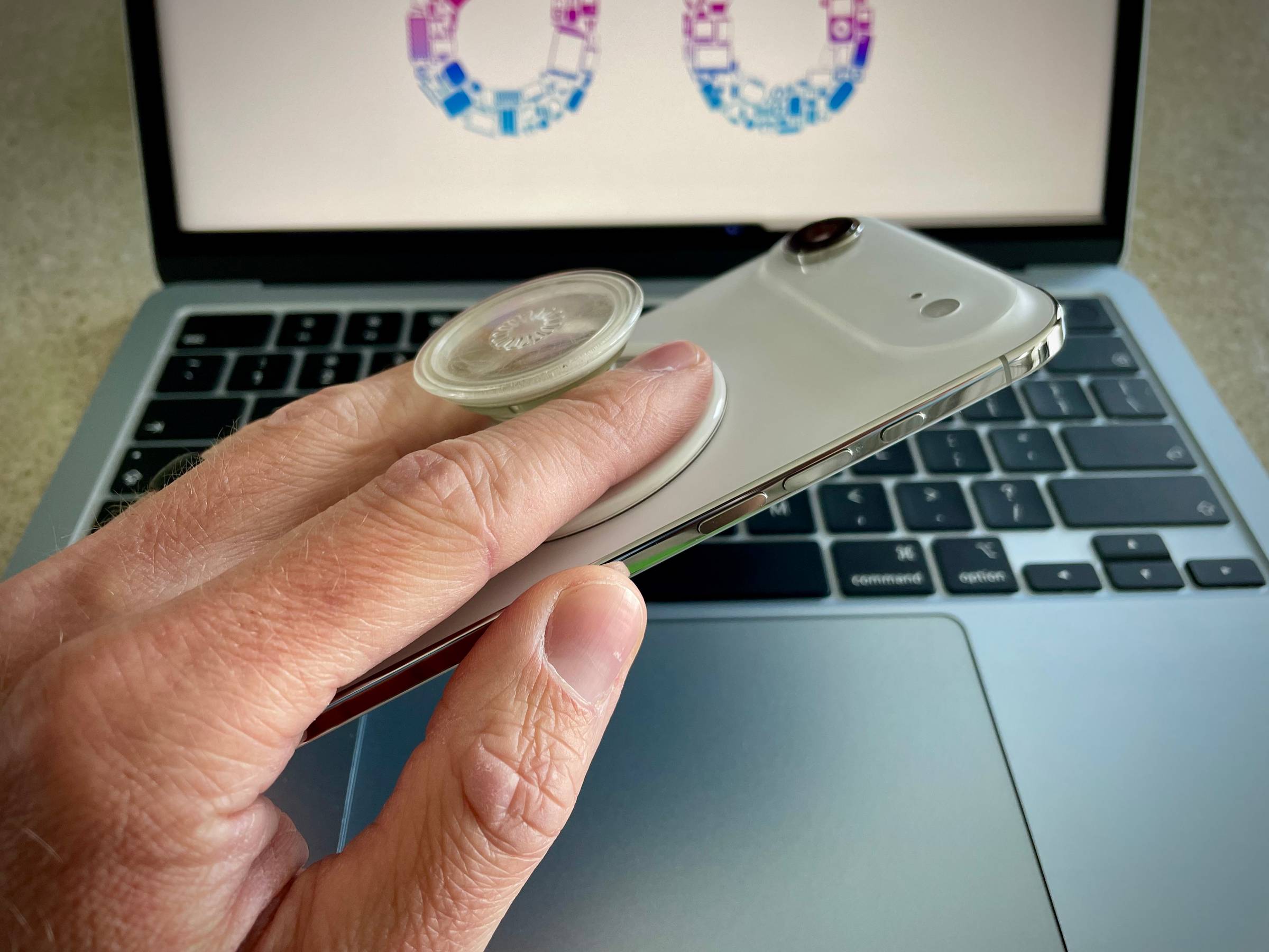 Hand holding an iPhone Air in a clear case at an angle over a MacBook keyboard. A clear PopSockets MagSafe grip is fully extended and popped open, with the thumb looped through the grip to demonstrate one-handed hold on a large phone.