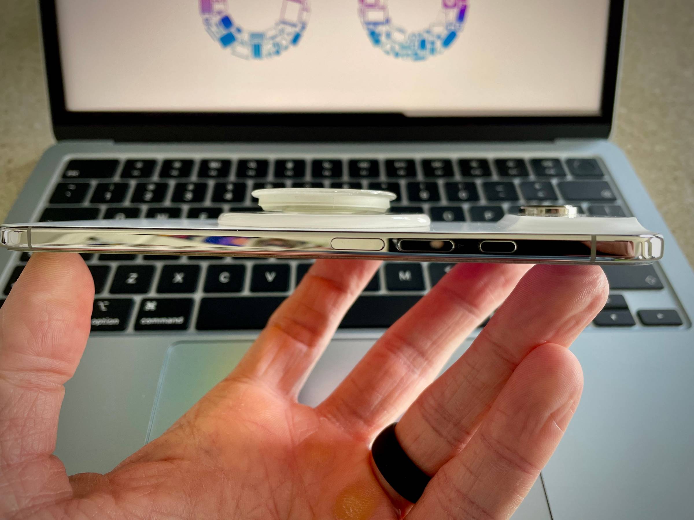 Side view of an iPhone Air in a clear case held flat in one hand above a MacBook keyboard. A clear PopSockets MagSafe grip is collapsed flat against the back of the phone, showing how low-profile it sits when not in use.