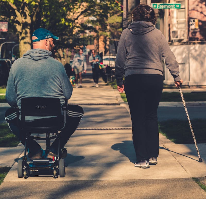 2 people walking, one using a cane, and the other riding in an electronic wheelchair
