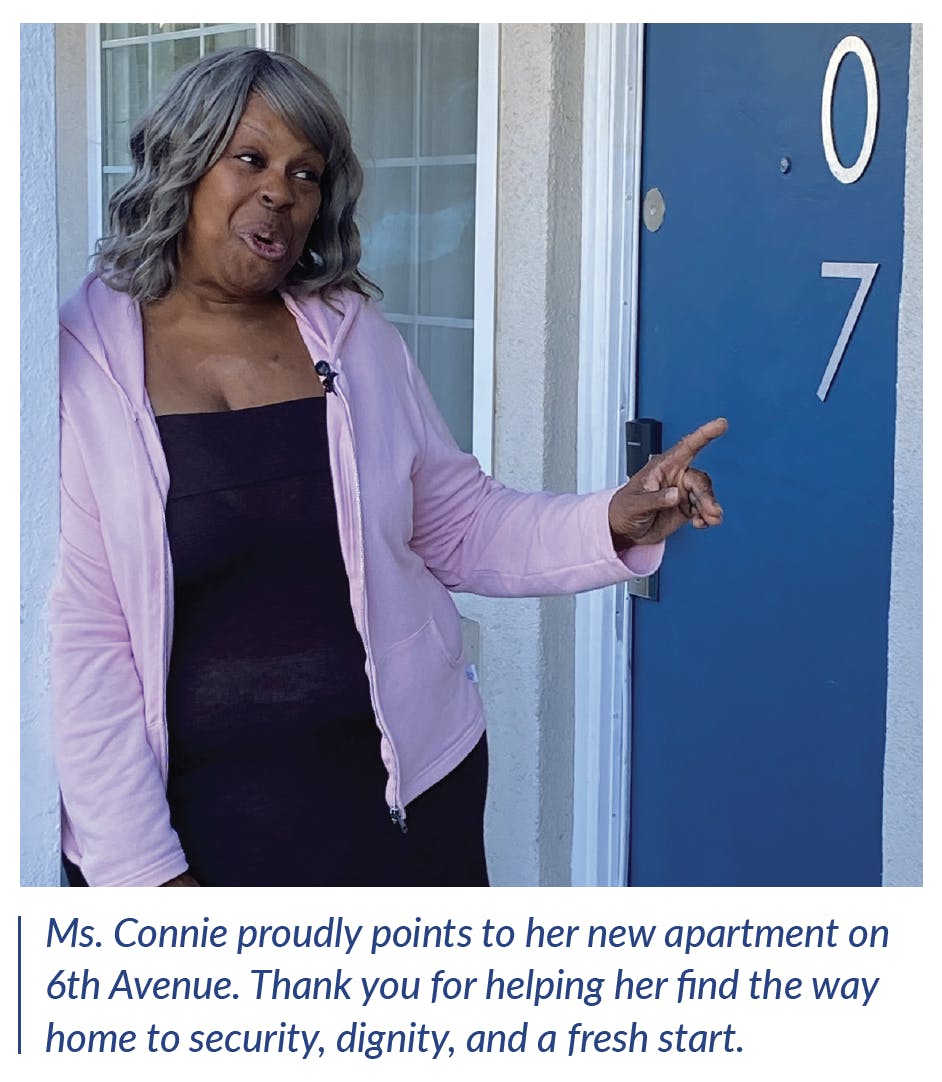 A woman pointing to her apartment door. The caption reads: Ms. Connie proudly points to her new apartment on 6th Avenue. Thank you for helping her find the way home to security, dignity, and a fresh start.