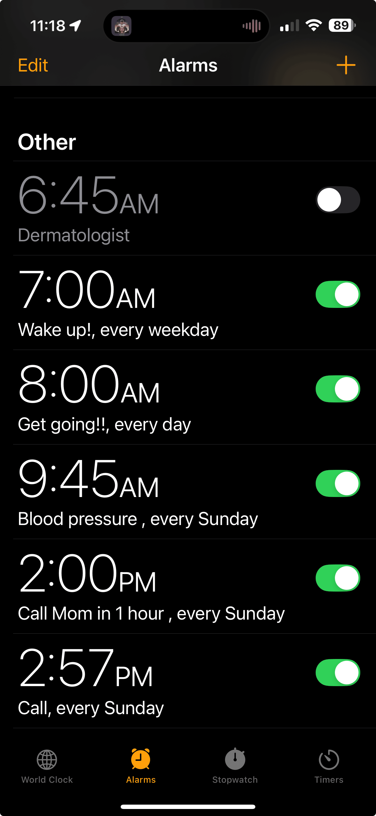 This is a screenshot of the Alarms tab in the iPhone Clock app. The screen is dark with white and orange text, showing a list of customized alarms under the section labeled “Other.” The time at the top of the screen is 11:18, and the battery is at 89%.  There are six alarms shown:  6:45 AM — Labeled “Dermatologist.” This alarm is switched off.  7:00 AM — Labeled “Wake up!, every weekday.” This alarm is on.  8:00 AM — Labeled “Get going!!, every day.” This alarm is on.  9:45 AM — Labeled “Blood pressure, every Sunday.” This alarm is on.  2:00 PM — Labeled “Call Mom in 1 hour, every Sunday.” This alarm is on.  2:57 PM — Labeled “Call, every Sunday.” This alarm is on.