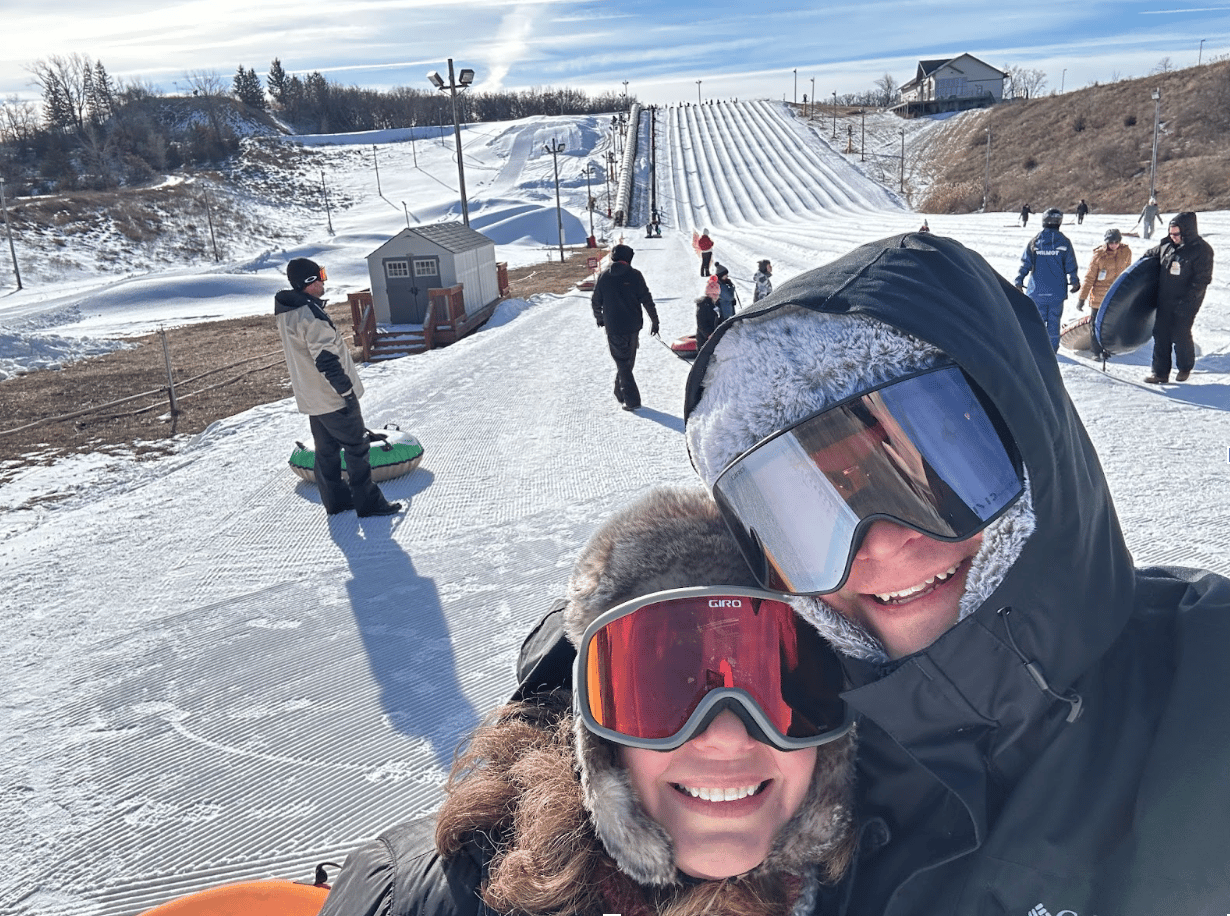 Brett and Julie in winter gear posing near a sledding hill