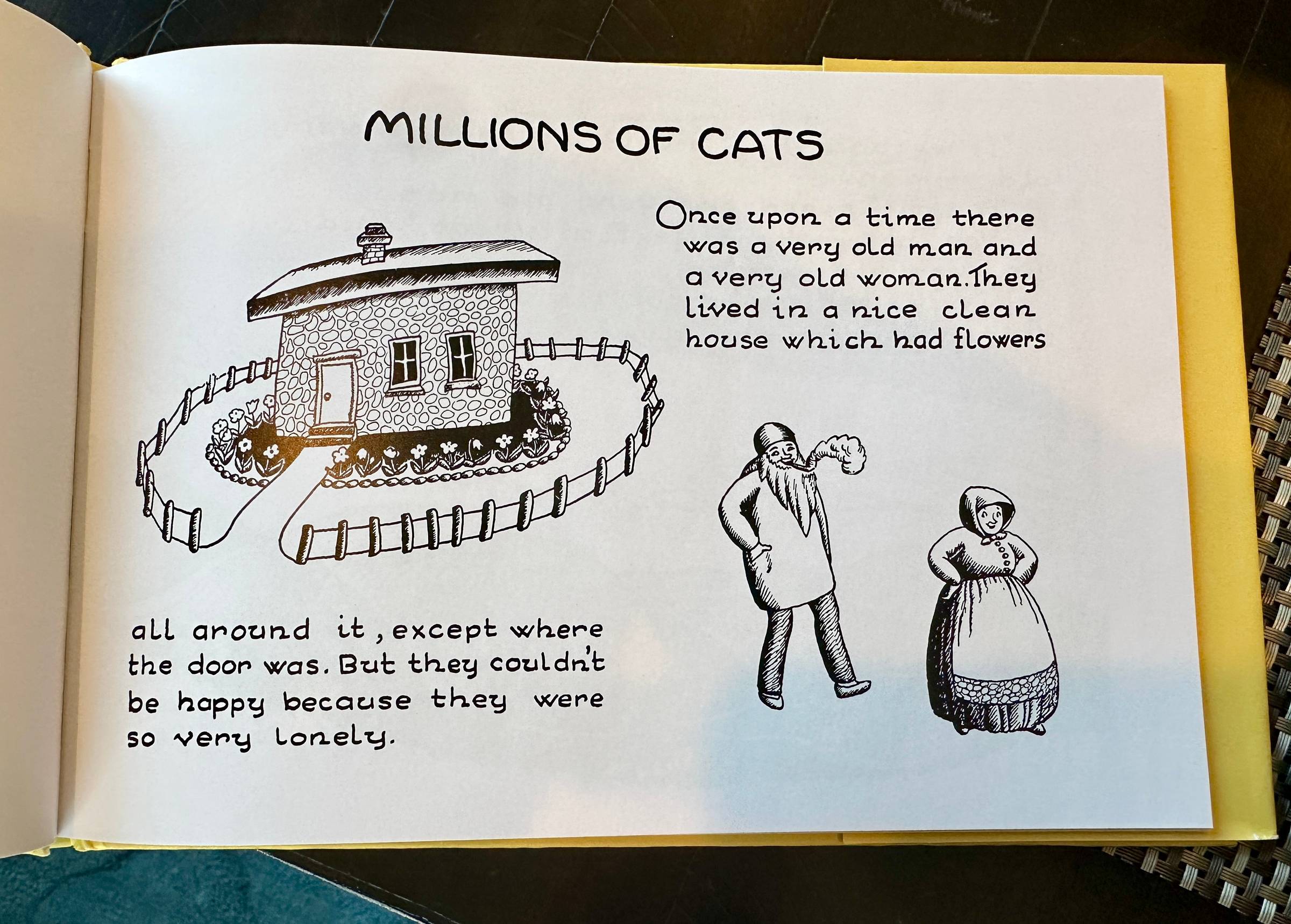 “An open children’s picture book titled ‘Millions of Cats.’ The left page shows a simple black-and-white drawing of a small stone house surrounded by a fence and flowers. The right page shows a very old man with a long beard and a very old woman standing 