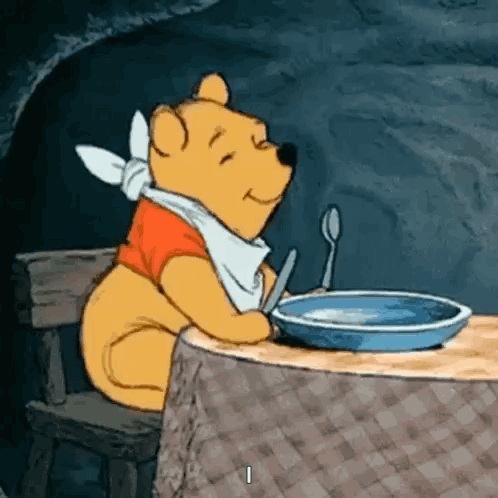 GIF of Winnie the Pooh sitting with utensils in hand before an empty plate, shifting side to side excitedly as he happily waits to be served a meal