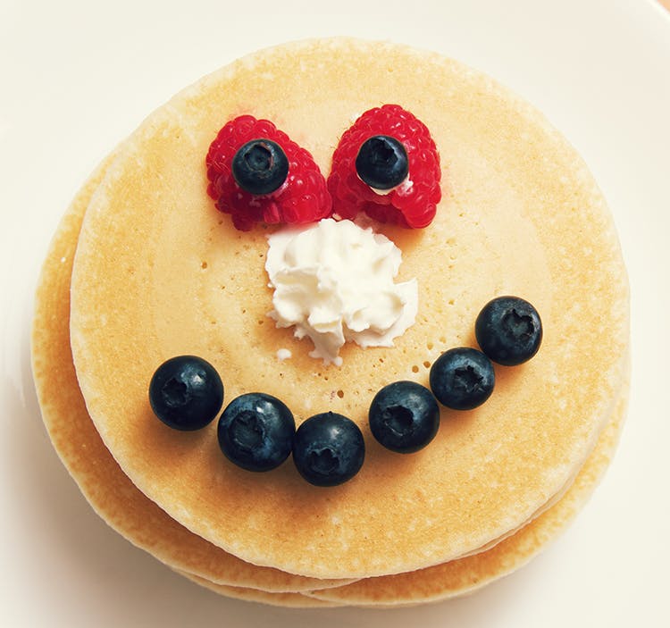 a stack of pancakes. the top one has a smiling face made of strawberries, blueberries and whipped cream