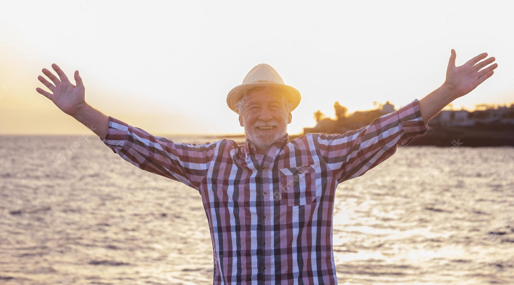 an elder man smiling with arms spread wide by the waterside in the magic or golden hour