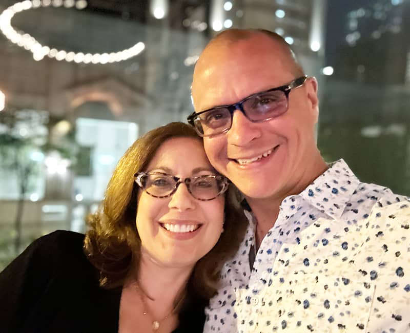 Julie Cooper and Brett Cooper looking at the camera, in the evening in front of a window in downtown Chicago