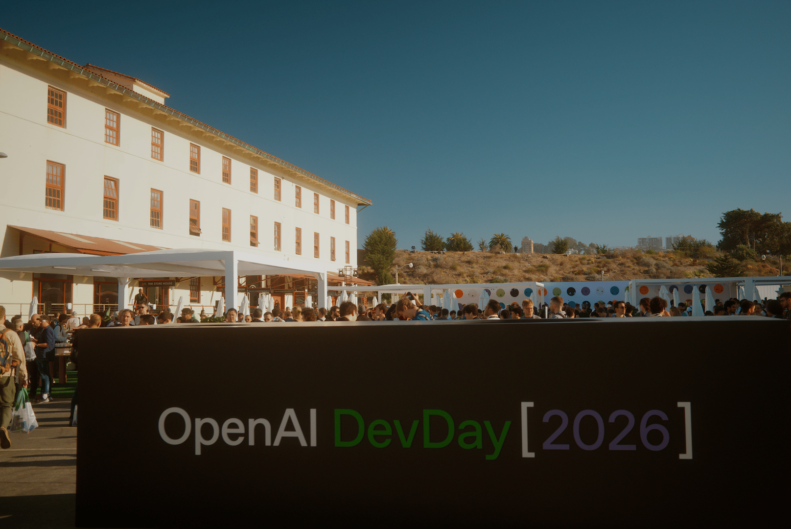 Outdoor reception at OpenAI DevDay in San Francisco, a crowd of people are behind a sign, next to an old building
