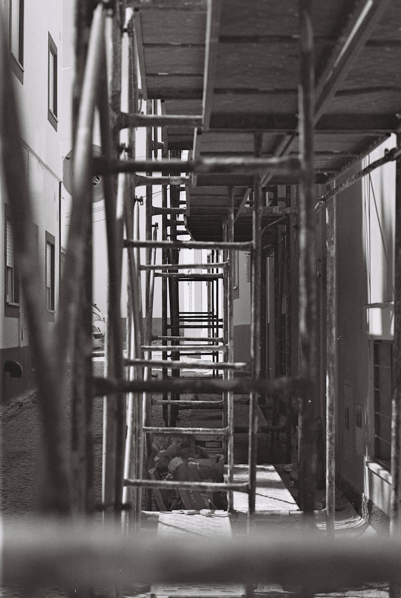 Metal scaffolding in a narrow alley between two whitewashed buildings in black and white