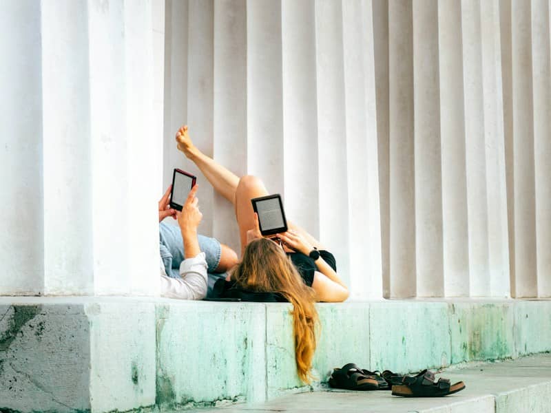 Two people reading e-readers while reclining against white fluted columns on a stone ledge