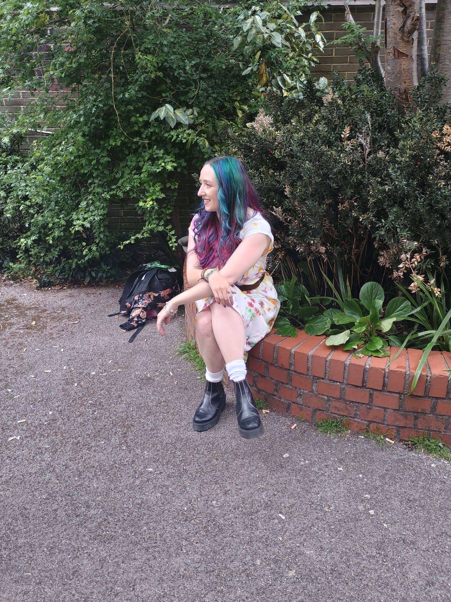 Beth sitting on a brick wall with arms learning on crossed legs, from a side perspective as she smiles towards a photographer to the left of the frame.