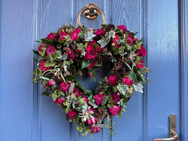 A heart shaped wreath made with roses and fresh flowers hung on a blue door