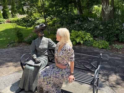 April Harris sitting beside a statue of L M Montgomery in the garden of Leaskdale Manse