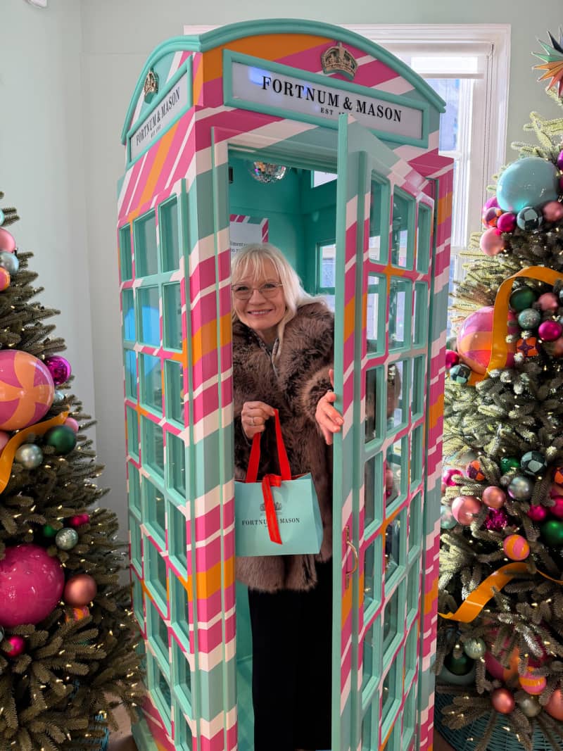 April Harris in a pastel coloured striped phone box at Fortnum and Mason in London. She is wearing a faux fur coat and holding a Fortnums bag. 