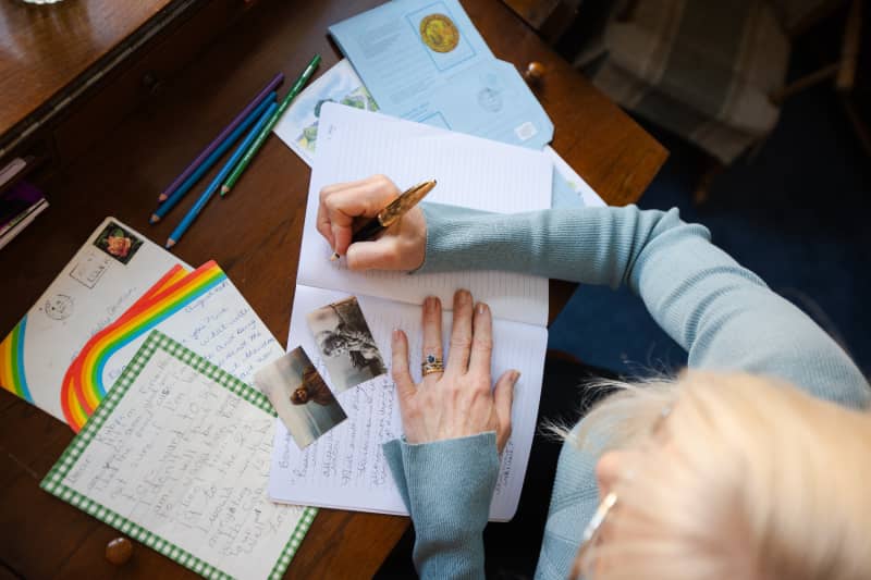 A photo taken from above of a woman writing in her diary. There are two small pictures of her as a younger woman and a child and some old letters in shot as well. 