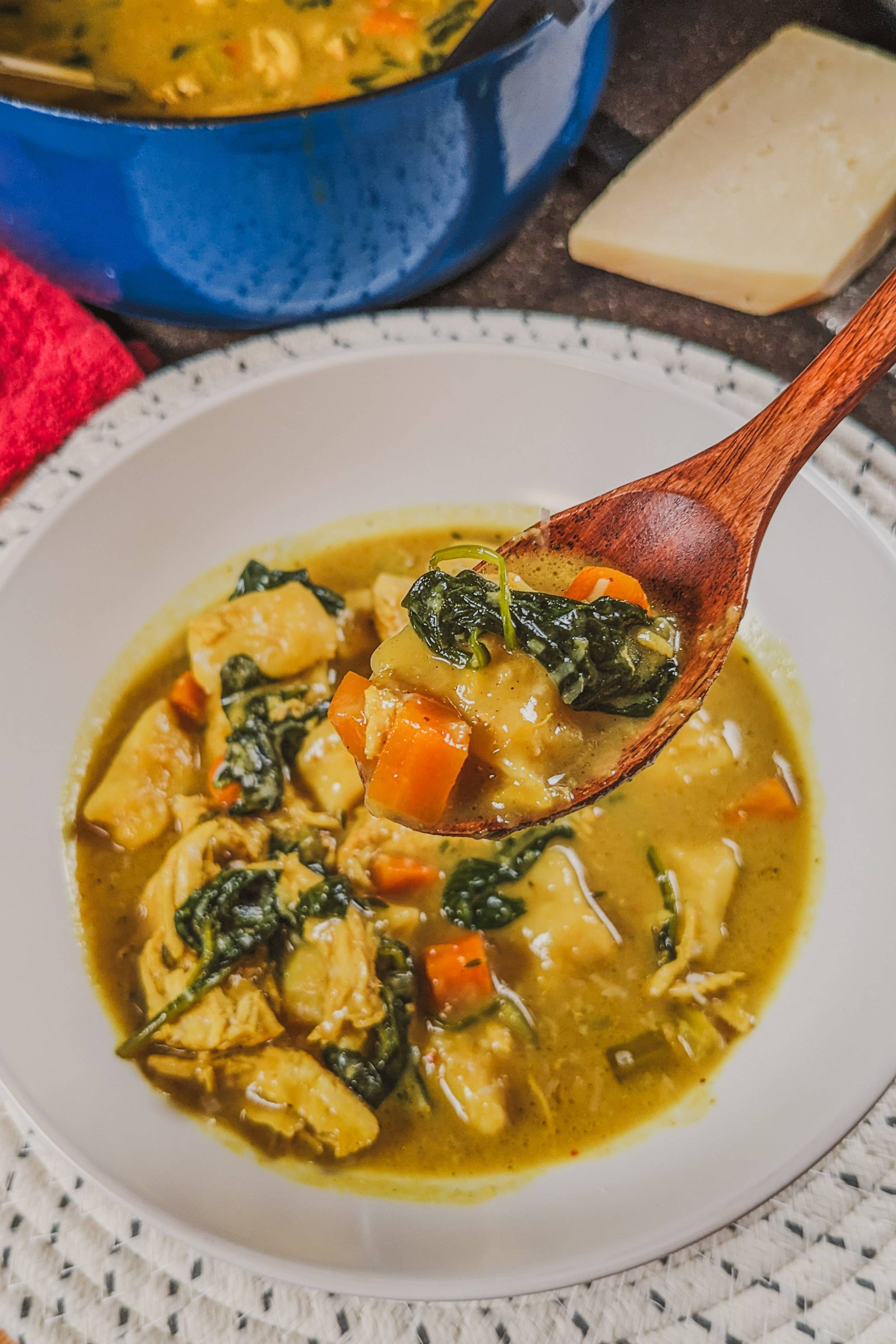 creamy curry chicken gnocchi soup with spinach and chunks of carrot and shredded chicken and large hand cut gnocchi sprinkled with freshly grated parmesan cheese in a wide rim white bowl with a wooden spoon.