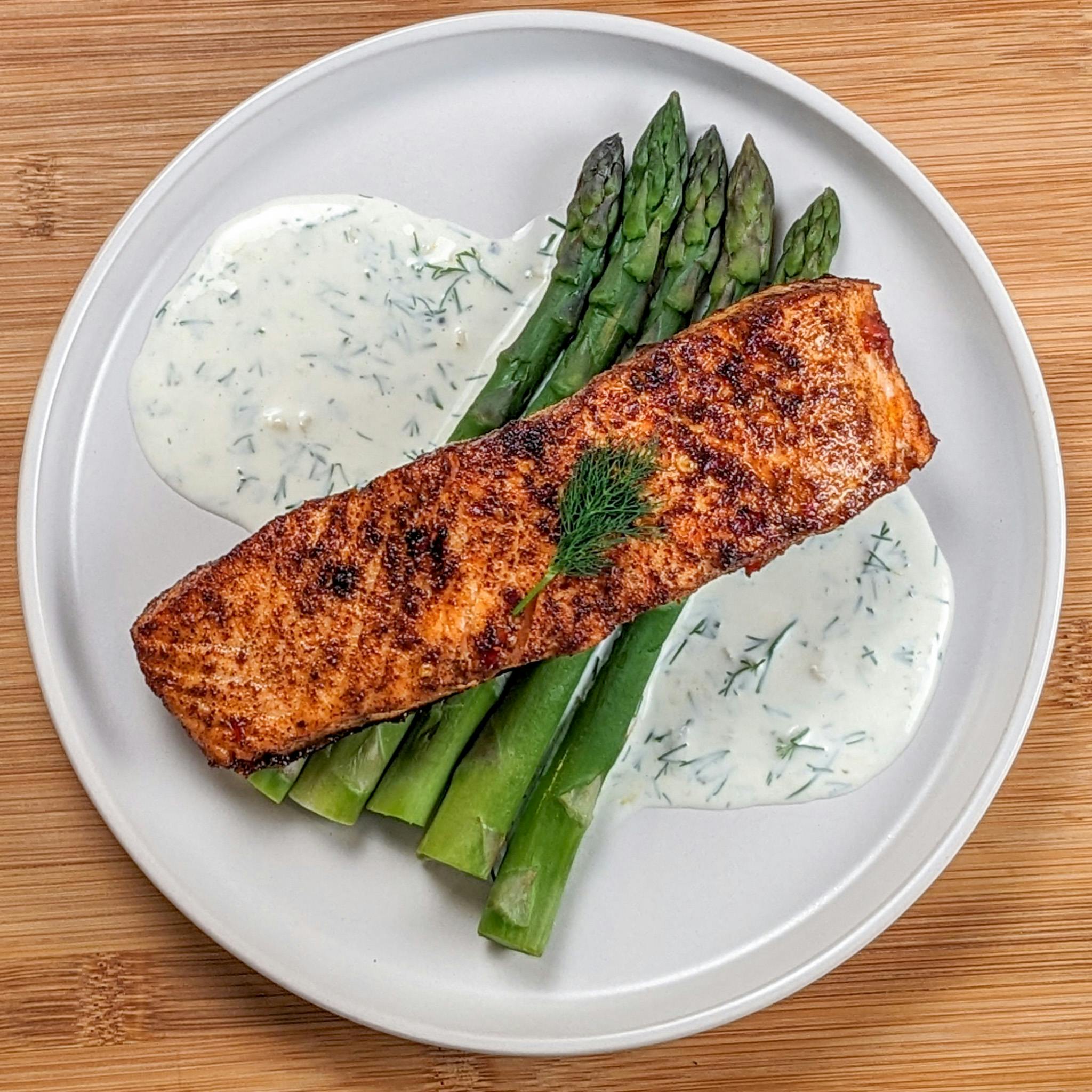 pan seared salmon on steamed jumbo asparagus on lemon dill sauce on a round flat plate.