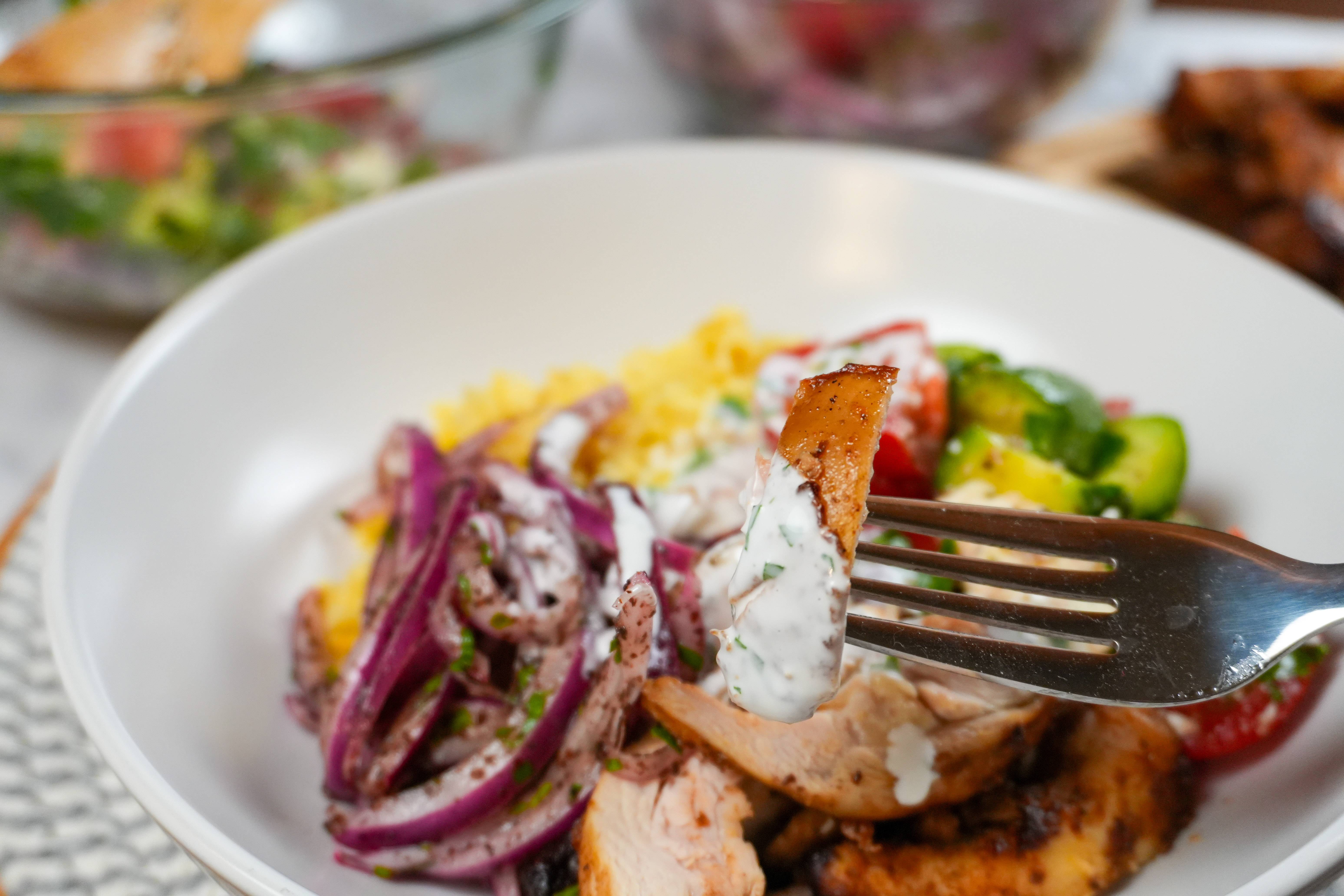 a fork of cooked chicken drizzled with lemon greek yogurt dressing with a middle easter inspired rice bowl in the background.