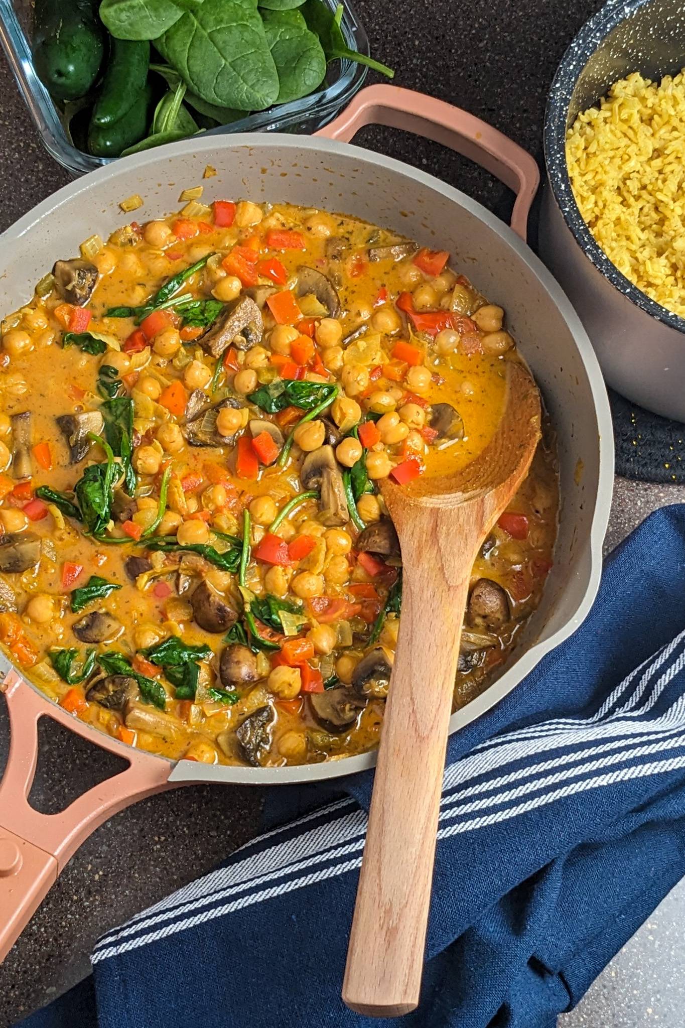 curried chickpeas with tomatoes, spinach and mushrooms in a skillet with a wooden spoon next to a kitchen towel and a container of spiced basmati rice.