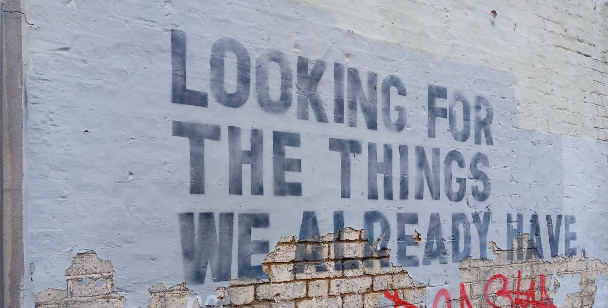 Image of graffiti on a wall.  Grey text on white paint.  'Looking for the things we already have'