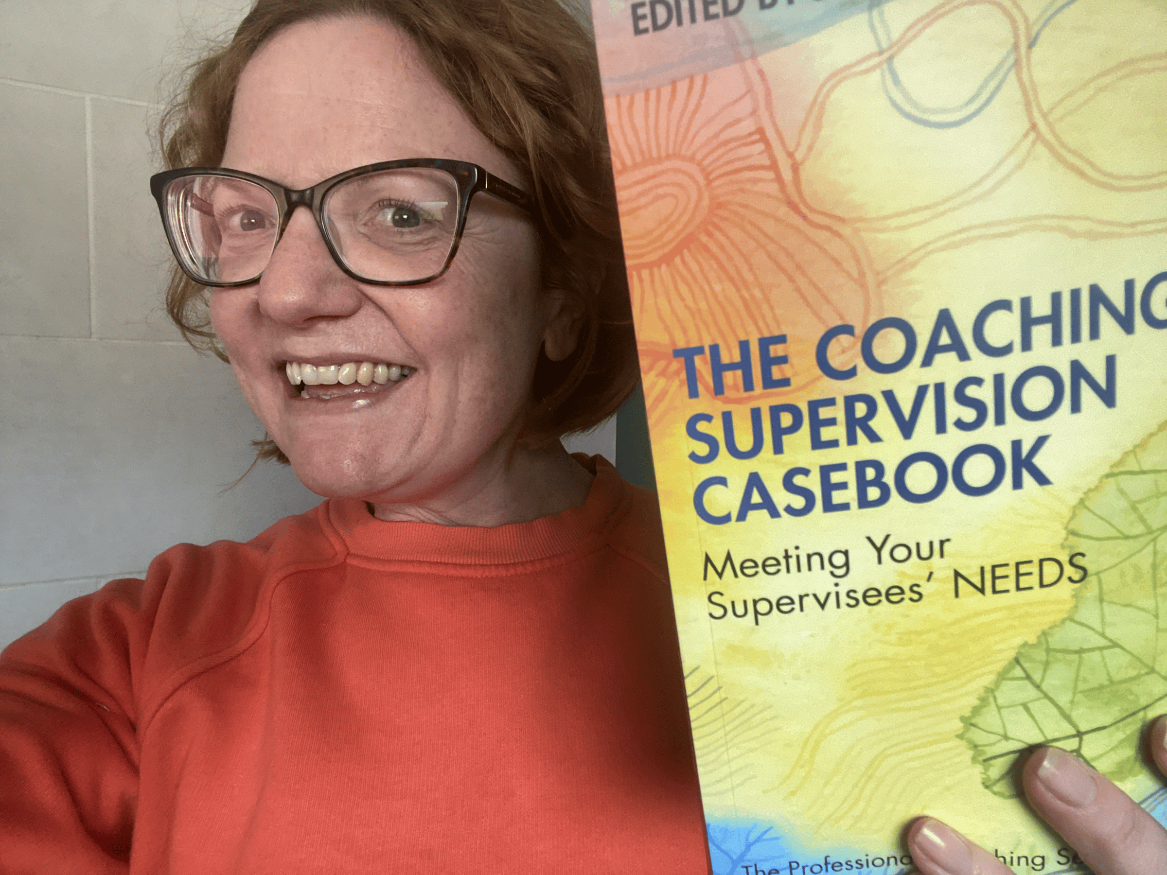 Sheela Hobden in an organge jumper, holding a copy of the book 'The Coaching Supervision Casebook:  Meeting Your Supervisees' NEEDS' edited by Jeremy Lewis, in which Sheela has a published chapter.