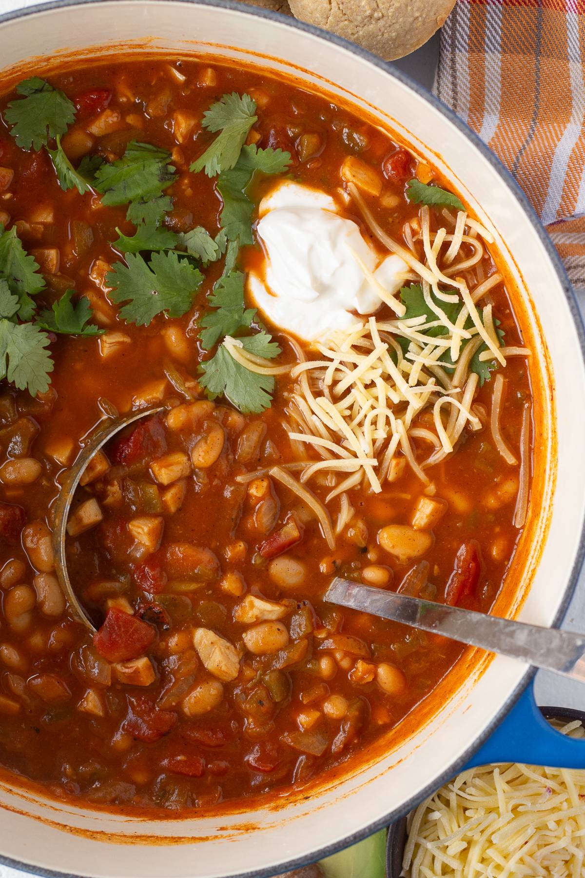 Top down shot of a pumpkin turkey chili in a large pot with cilantro, sour cream, and cheese on top, with a silver ladle in it.